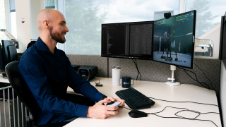 Man in office cubicle holds white controller, testing UFC 4 on dual monitors showing code and gameplay, with EA Sports mug and gaming setup on desk