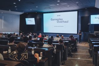 Publikum in einem Konferenzraum, das eine "Gameplay-Überarbeitung"-Präsentation von Sam Rivera und Paul Parsons auf einem großen Bildschirm verfolgt.