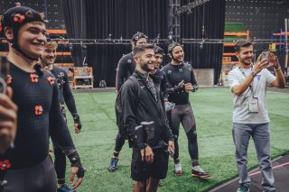 Eine Gruppe von Menschen in Motion-Capture-Anzügen versammelt sich auf einem grünen Kunstrasenfeld in einem Studio-Setting, umgeben von Ausrüstungen und Gerüsten.