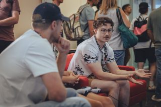 Eine Gruppe von Menschen versammelt sich in entspannter Atmosphäre. Zwei Personen sitzen auf einer roten Couch und unterhalten sich. Andere stehen im Hintergrund und plaudern.