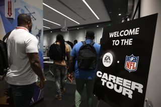 People with backpacks enter a room for the EA Sports and NFL Career Tour, as a sign welcomes them. The room has modern decor with a projector and screen.