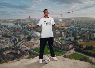 Grafica di EA SPORTS FC™ 27 che mostra Kylian Mbappé, con la divisa del Real Madrid, in piedi a braccia aperte su un pallone da calcio che sovrasta un panorama urbano ricco di stadi, bandiere dei club e monumenti simbolo.