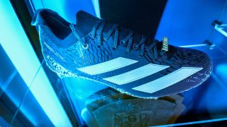 Close-up of a black and blue Adidas football cleat with white stripes displayed in a lit glass case, with sports memorabilia visible in the background.