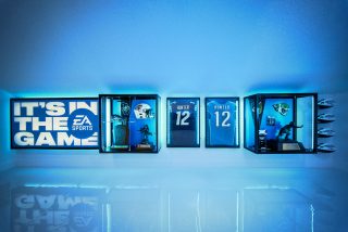 EA Sports memorabilia wall display with glowing blue light, showcasing framed football jerseys, a helmet, trophies, and a large 'Its™ In The Game' sign.