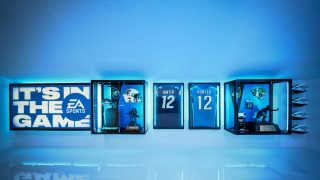 EA Sports memorabilia wall display with glowing blue light, showcasing framed football jerseys, a helmet, trophies, and a large 'Its™ In The Game' sign.