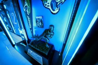 Close-up of a bronze football player trophy in a lit display case, surrounded by Jacksonville Jaguars memorabilia and an EA Sports College Football 25 game cover.