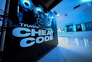 Close-up of a custom gaming PC with glowing blue lights and graphics featuring 'Travis Hunter Cheat Code' text, with EA Sports memorabilia wall in the background.