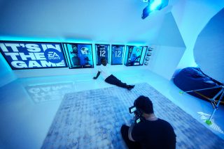 A photographer captures Travis Hunter in a white shirt sitting on the floor in front of EA Sports-themed memorabilia and framed football jerseys glowing with blue lighting.