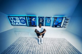 Travis Hunter sits on a carpet in a bright blue-lit room, with framed football jerseys, helmets, and EA Sports memorabilia displayed on the wall behind him.