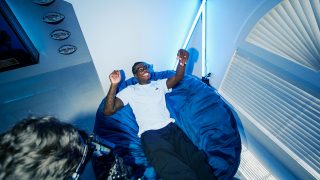 Travis Hunter in a white shirt and black pants reclines on a large blue beanbag, smiling as a cameraman films him in a room lit with bright blue neon lights.