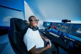 Travis Hunter in a white shirt and glasses sits in a gaming chair with headphones around his neck, holding a controller at a desk setup with three monitors and streaming gear.