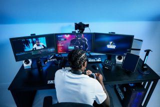 Travis Hunter in a white shirt sits in a gaming chair with headphones around his neck, holding a controller at a desk setup with three monitors and streaming gear.