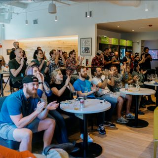 A group of people sitting and standing in a casual setting, clapping and watching something off-camera with interest and engagement.