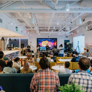 A lively office event with people gathered in a modern space, featuring a band performing on stage and a large screen displaying colorful graphics in the background.