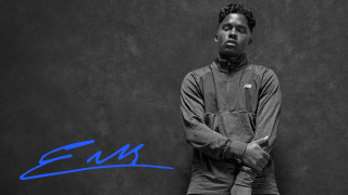 Black and white picture of Endrick, leaning back on a wall with his hand grabbing his other arm. His signature in blue is at the bottom left of the picture. 