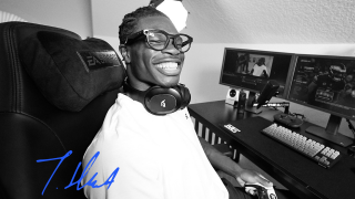 Travis Hunter in a white shirt and glasses sits in a gaming chair with headphones around his neck, holding a controller at a desk setup with three monitors and streaming gear. The picture is in black and white and his signature in blue is in the left bottom corner. 