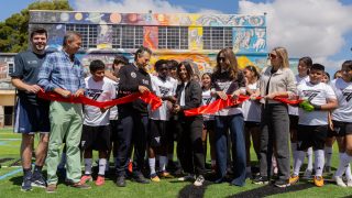Kids and their trainers in EA SPORTS FC t-shirts cutting a red ribbon in front of a school painted with vibrant colors. 