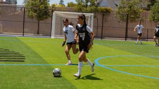 Two young girls in EA SPORTS FC training tops pursue a ball on a green pitch. 