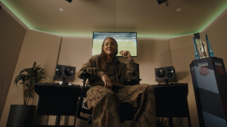 Alex Scott posing to the camera. Behind her there are a pair of speakers and a monitor playing a soccer match