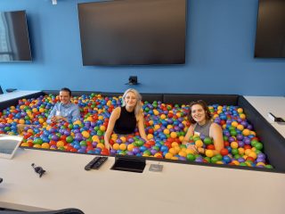 Three smiling people, two with name tags, are chest-deep in a multi-colored ball pit. The ball pit is in the center of a conference table with teleconference equipment on it. There are three large-screen TVs on the wall behind.