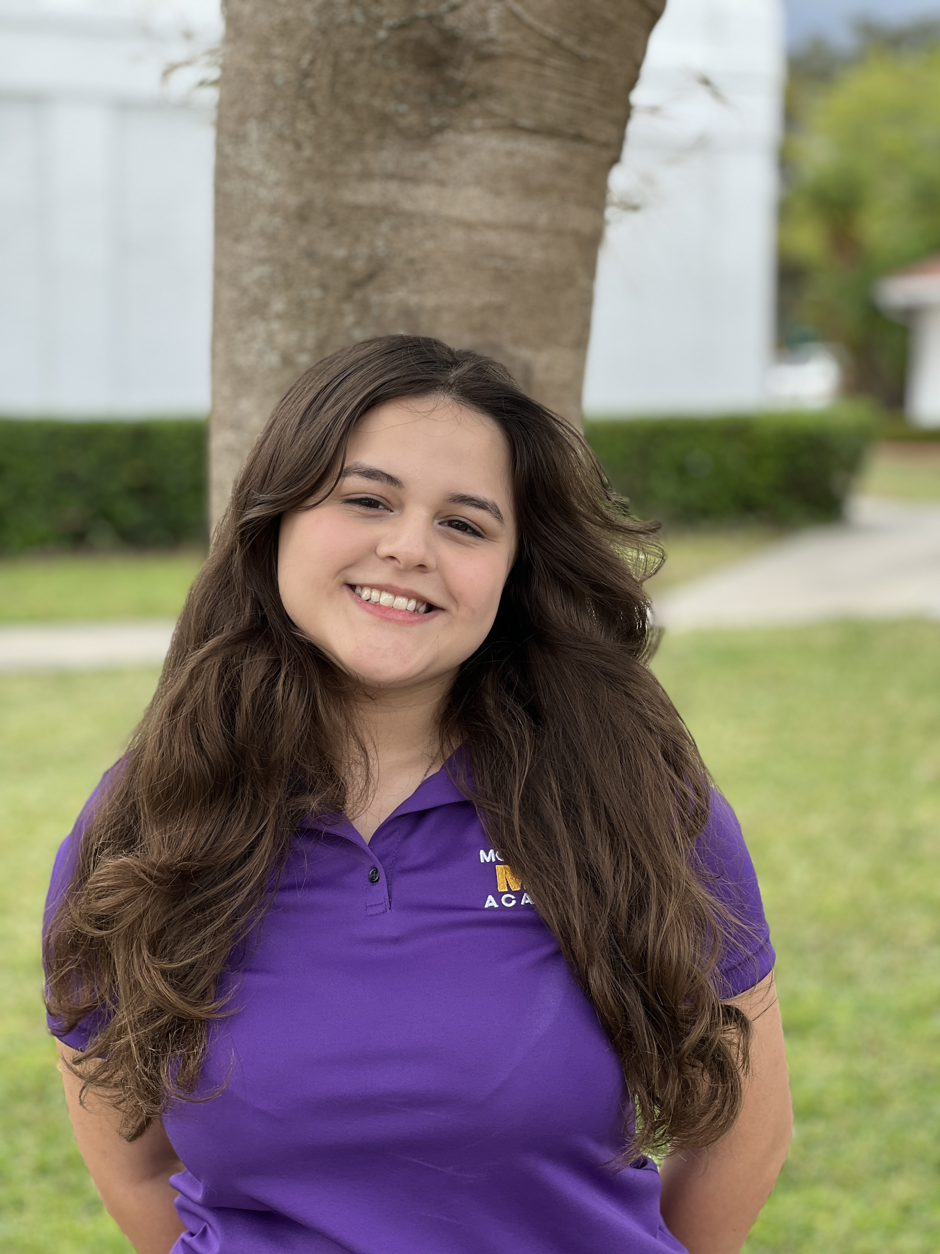 A young female student with brown hair and a purple polo shirt smiles at the camera while standing in front of a tree.