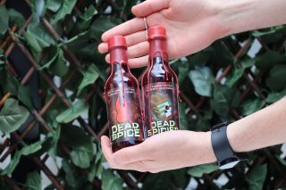 A person holds 2 bottles of Dead Space themed hot sauces, with one having Isaac Clarke's helmet, and the other having the marker.