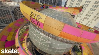 Skaters navigate a vibrant, twisting skatepark with pink and yellow ramps encircling a large glass dome in an urban setting with tall buildings.