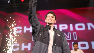 En leende man höjer armen på scen framför en röd bakgrund med texten ”CHAMPION” och gnisteffekter, för att fira en seger vid ett evenemang.