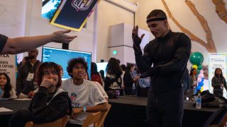 A man in a black motion capture suit demonstrates hand movements in front of two seated youths wearing NFL shirts, with onlookers and NFL signs in a busy event space.