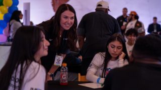 A woman wearing a "Culture Club Guest" badge smiles and leans toward a table where young people, one with an NFL shirt, sit and write. Balloons are in the background.