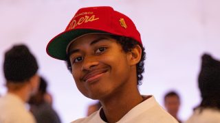 A young person smiles at the camera wearing a red San Francisco 49ers cap and a white shirt, with several people in the background.