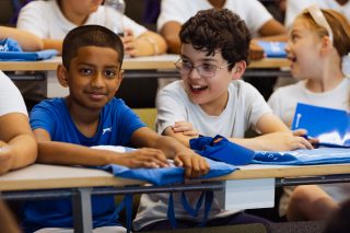 Children sit at desks in a classroom setting, engaged in an educational activity. They are wearing casual clothing, and some hold blue bags, suggesting a workshop or event.