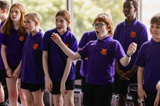 A group of children wearing matching purple polo shirts with school emblems stand in a classroom. They appear engaged and animated, participating in an interactive activity.