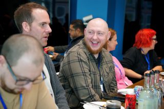 A group of people seated at a conference table with blue lanyards, engaged in discussion. The table is set with drinks and bottles, and a person with bright red hair is visible.