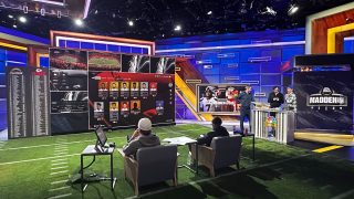 EA Sports Madden NFL studio with a game on and many screens showing, stats, players, and more. Two people are seated watching the data while three stand near a presentation stand having a conversation.