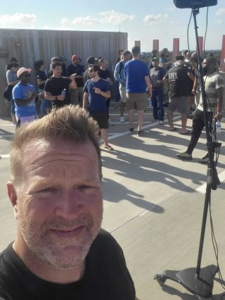 A group of people gathered on a rooftop under a clear sky, engaging in conversation and enjoying a sunny day. Shadows are cast on the concrete surface.