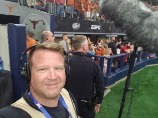 A crowded sports stadium with fans in burnt orange attire, a police officer standing near the field, and a large boom microphone capturing the event's sounds.