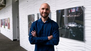 Smiling bald man with beard stands in modern office hallway wearing dark blue shirt, arms crossed, with framed UFC arena photo and artwork on white wall behind him.