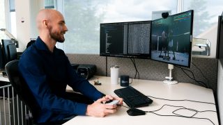 Man in office cubicle holds white controller, testing UFC 4 on dual monitors showing code and gameplay, with EA Sports mug and gaming setup on desk